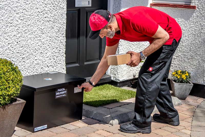 Photograph showing a courier with a parcel in their hand, pressing the button on the front of the iParcelBox smart parcel delivery box to request access to deliver a parcel.