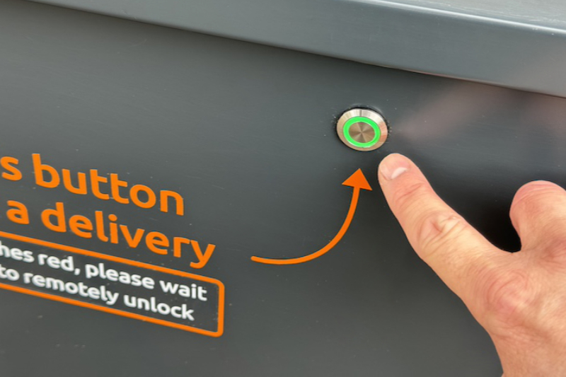 Photo showing courier pressing the button to request a parcel delivery. The light around the button is green, indicating that the parcel drop box is unlocked and ready to accept a parcel delivery.